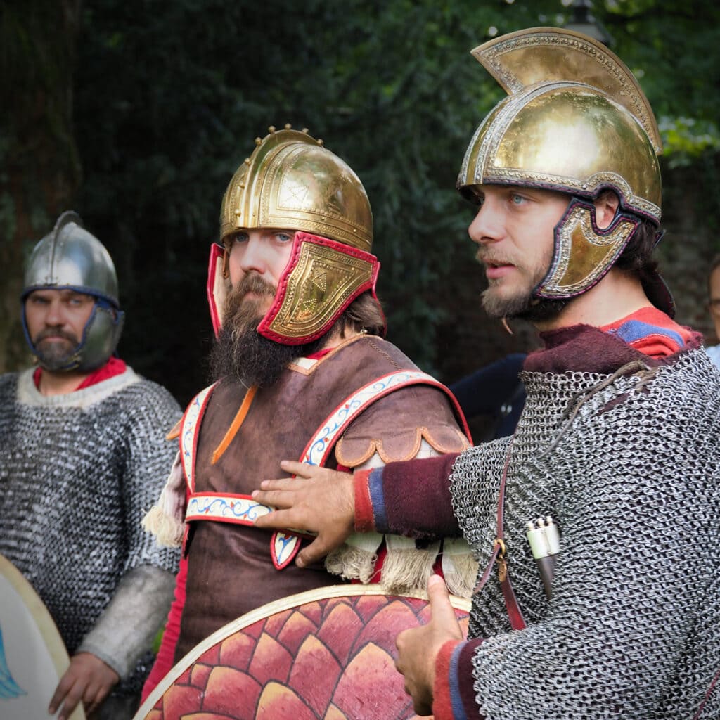 Drei Männer in historischen Rüstungen mit Kettenhemden und glitzernden Helmen, ernste Gesichter.