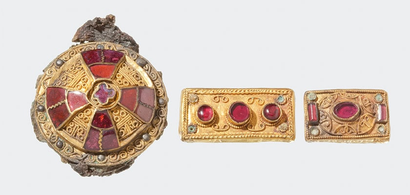 Drei antike goldene Schmuckstücke mit roten Edelsteinen und filigranen Verzierungen auf neutralem Hintergrund.