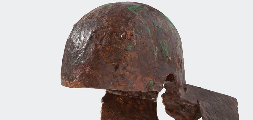 Rostiger antiker Metallhelm, seitliche Ansicht, leicht beschädigt, braun mit grünen Patina-Flecken.