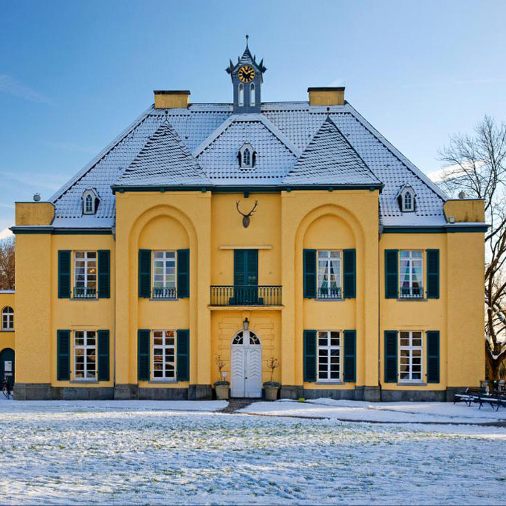 Gelbes Herrenhaus mit grünen Fensterläden und Uhrturm, verschneite Umgebung, blauer Himmel.