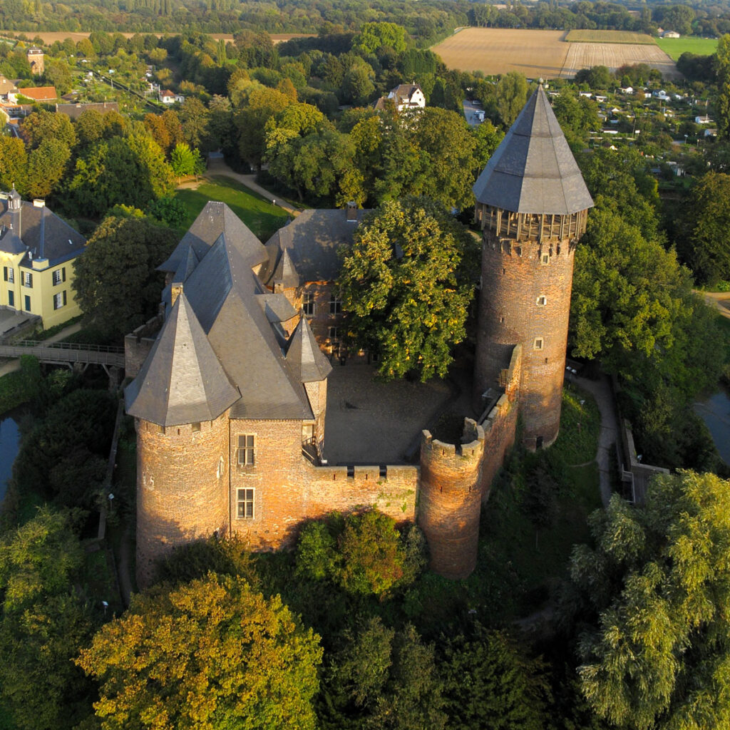 Luftaufnahme einer mittelalterlichen Burg mit Türmen, umgeben von Bäumen und ländlicher Landschaft.