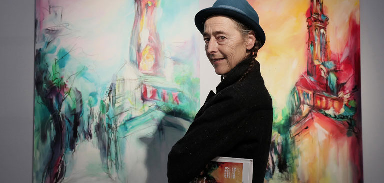 Person mit blauem Hut lächelt; hält ein Buch vor farbenfroher, abstrakter Kunst.