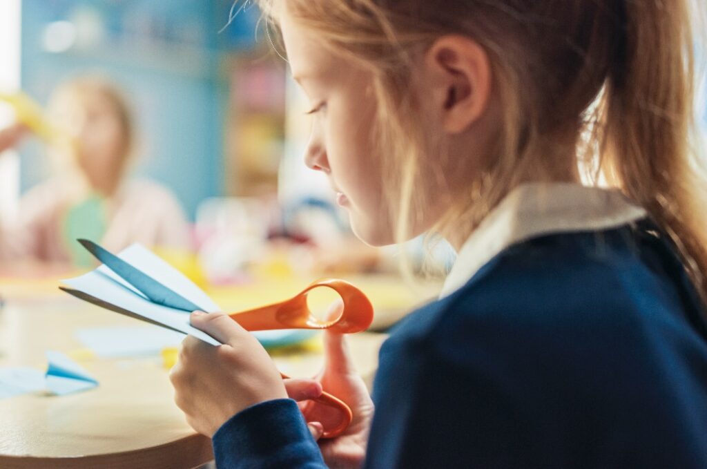 Grundschule Kunst & Handarbeitsklasse: Über die Schulter eines Mädchens hat Spaß am Bastelprojekt, schneidet Papierfiguren mit der Schere aus. Kreatives junges Mädchen
