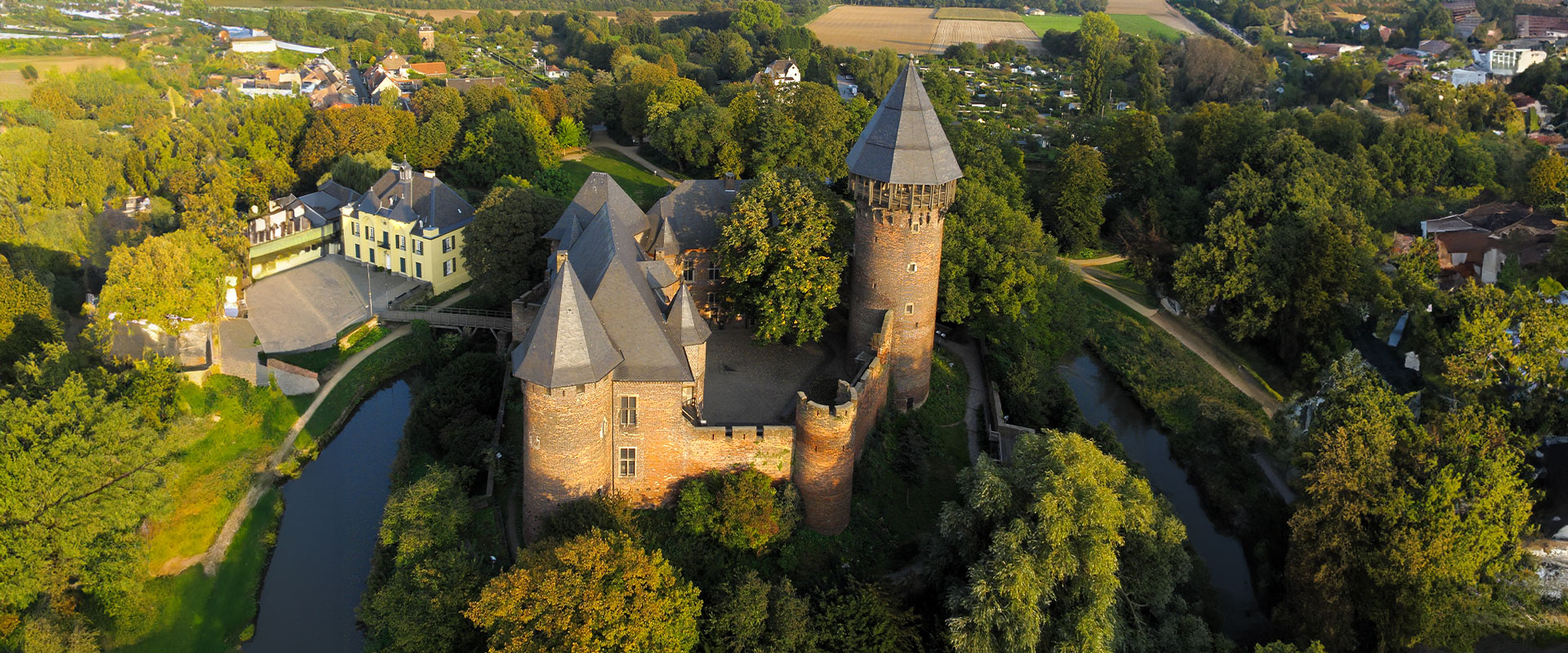 Luftaufnahme einer historischen Burg mit roten Türmen, umgeben von Bäumen und Wassergraben.
