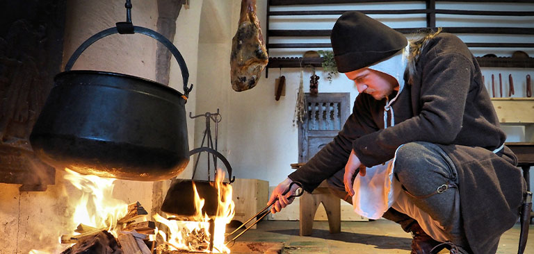 Ein Mann in mittelalterlicher Kleidung kocht am offenen Feuer, fokussiert, in einer rustikalen Küche.