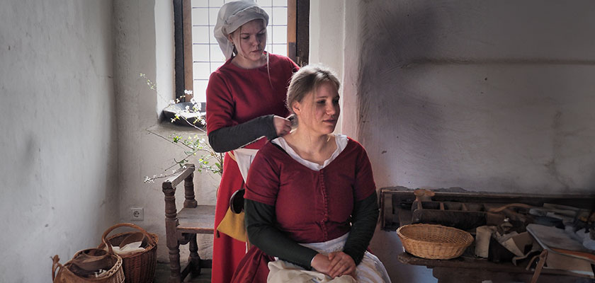 Zwei Frauen in mittelalterlicher Kleidung; eine flechtet der anderen das Haar; in rustikalem Raum.