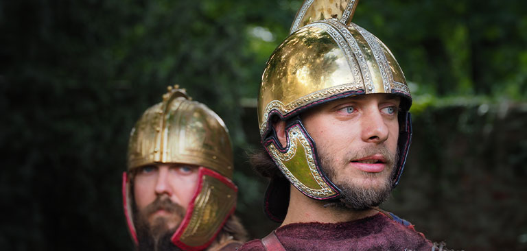 Zwei Männer in römischen Helmen, vorne mit nachdenklichem Blick, goldene Helme im Wald.