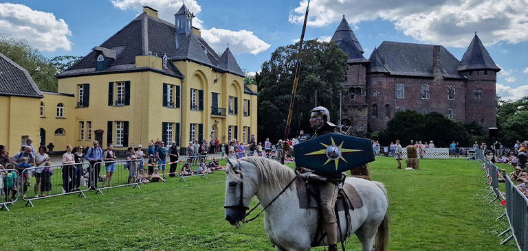 Ein Ritter in Rüstung auf weißem Pferd vor einer gelben Burg bei einem Mittelalterfest.
