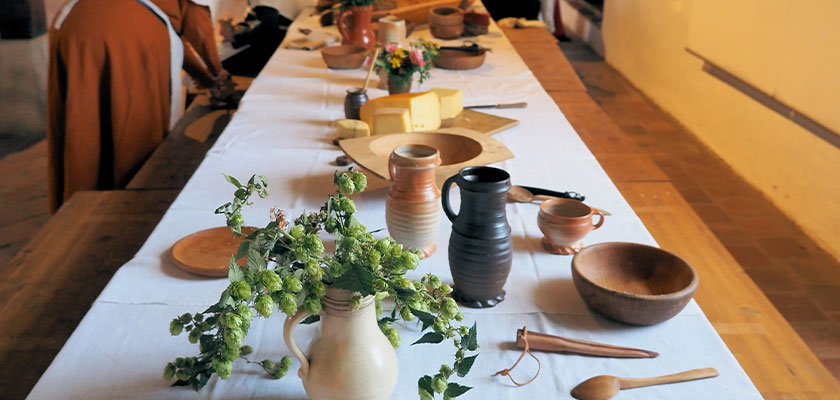 Langes Tischgedeck mit Tonkrügen, Holztellern und Blumenarrangement in historischem Ambiente.