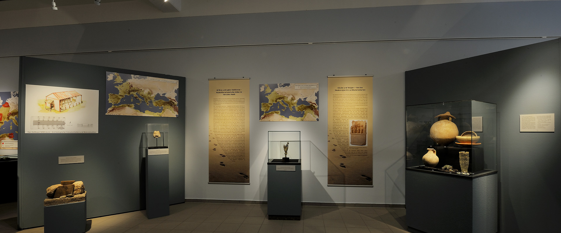 Ein Raum mit Ausstellungsstücken in Vitrinen, Texttafeln und Landkarten an grauen Wänden.