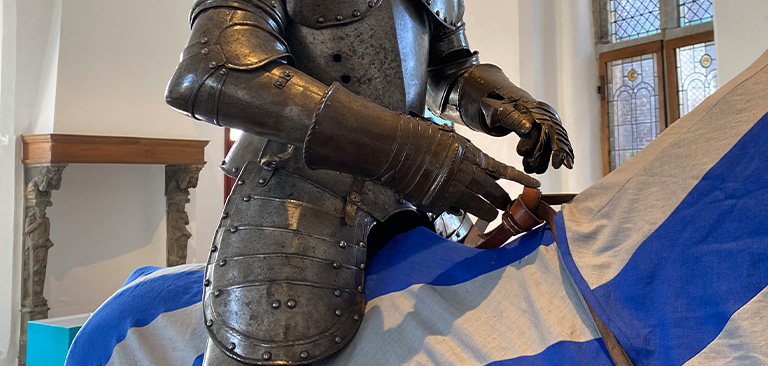 Ein Ritter in voller Rüstung deutet auf etwas, vor blauen und weißen Stoffen.