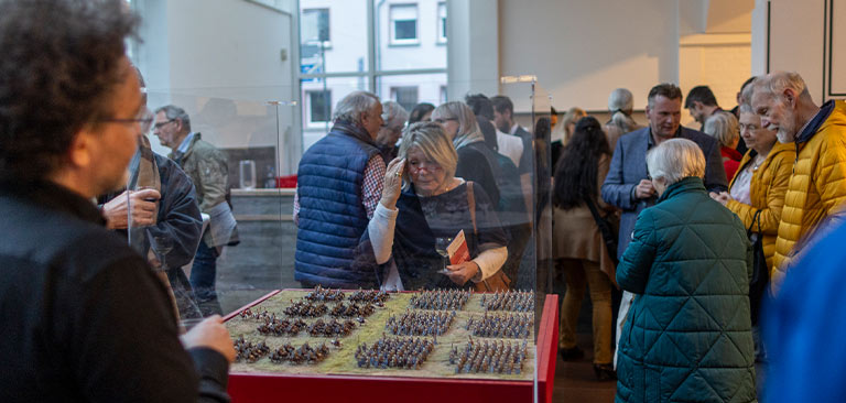 Gruppe von Personen betrachtet ein Miniaturmodell in einem Museum, viele tragen bunte Jacken.