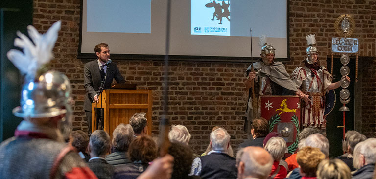 Eine Präsentation mit zwei römischen Soldaten in Uniform, Redner am Podium, Publikum fokussiert.