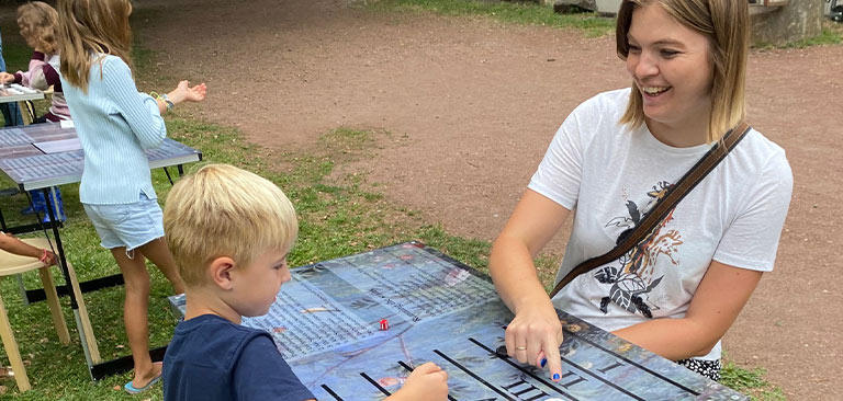 Eine junge Frau lächelt, während sie mit einem blondhaarigen Jungen an einem Tischspiel spielt.