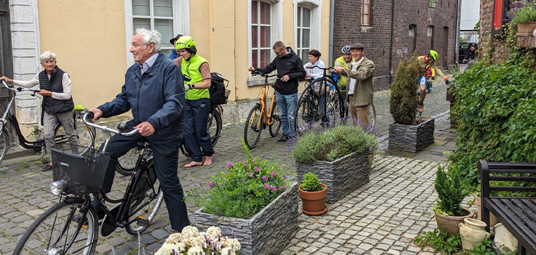 Ältere Person mit Fahrrad, lächelnd, in blauer Kleidung, andere Radfahrer im Hintergrund, Stadtgasse.