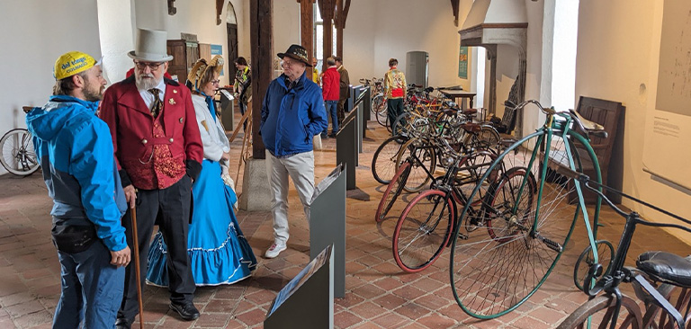 Personen in historischer Kleidung, diskutierend, mit alten Fahrrädern in einer Ausstellung.
