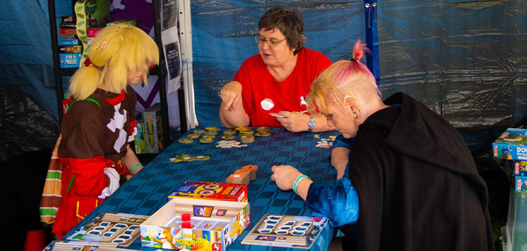 Drei Personen spielen ein Brettspiel; zwei sind verkleidet, eine erklärt. Buntes Spielzubehör.