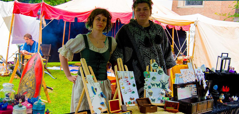 Zwei Personen bei einem Marktstand mit Kunsthandwerk, lächelnd, historische Kleidung, bunte Zelte.