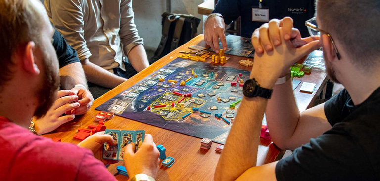 Personen spielen konzentriert ein Brettspiel an einem Holztisch mit bunten Spielfiguren.