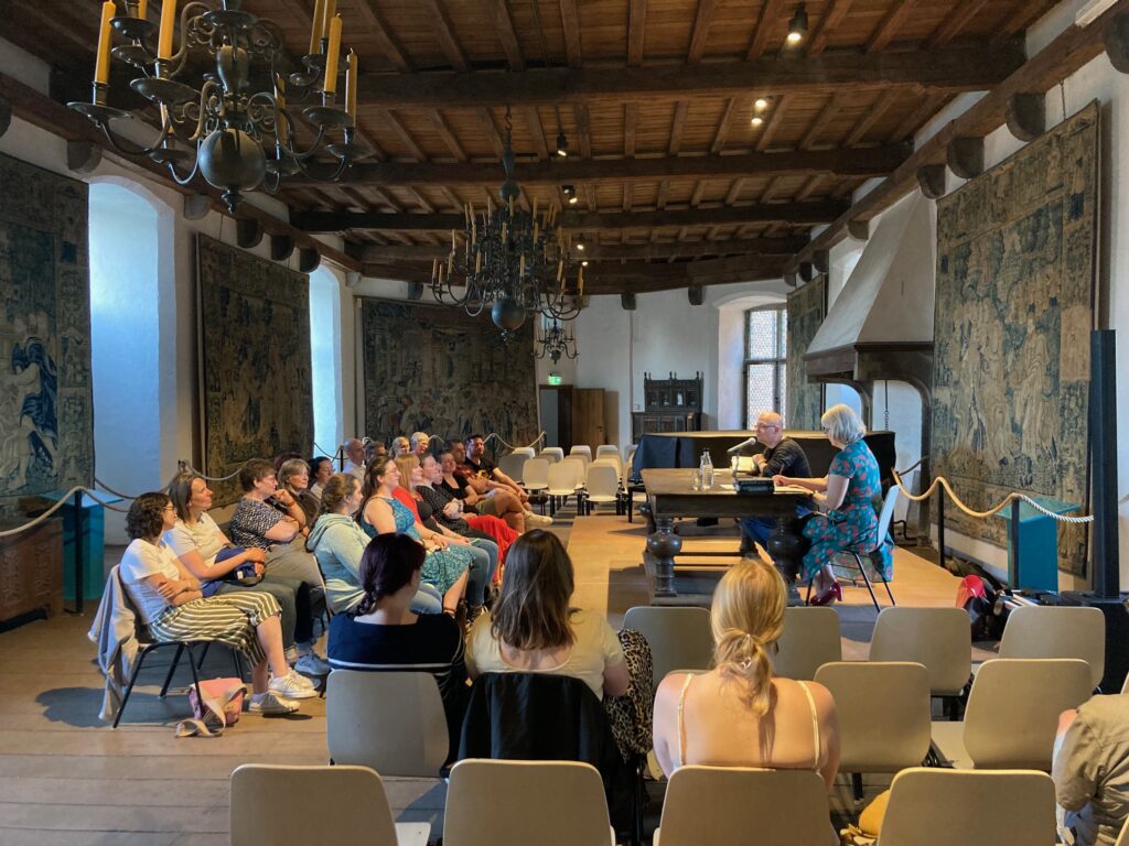 Eine Gruppe von Menschen in einem Saal mit Holzdach und Wandteppichen hört einem Vortrag zu.