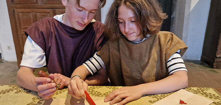 Zwei Jugendliche in historischen Gewändern schreiben zusammen an einem Tisch, konzentrierter Ausdruck.