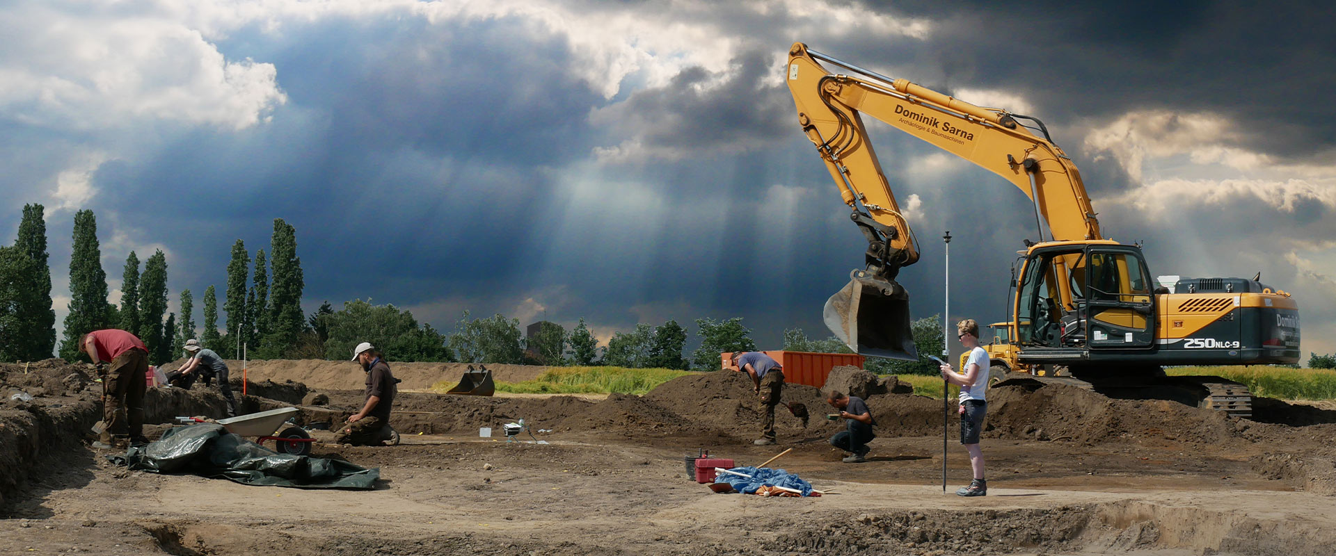 Mehrere Personen arbeiten mit Werkzeugen auf einer Baustelle bei bewölktem Himmel mit Bagger.