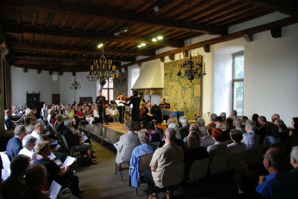 Ein Konzert im rustikalen Saal mit Holzdecke und Wandteppichen, gut besuchtes Publikum.