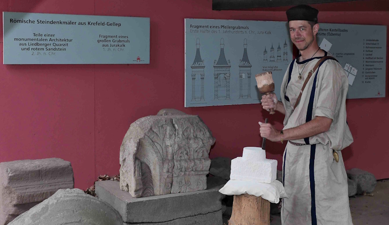 Laechelnder Mann in heller Tunika mit Hammer vor roemischen Steinen